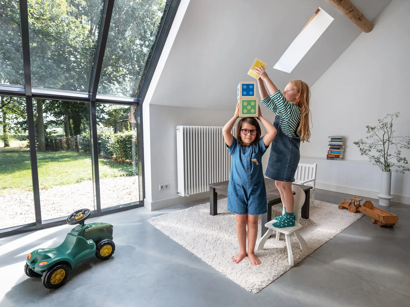 Twee kinderen stapelen houten blokken in een lichte zolderkamer met uitzicht op de tuin.