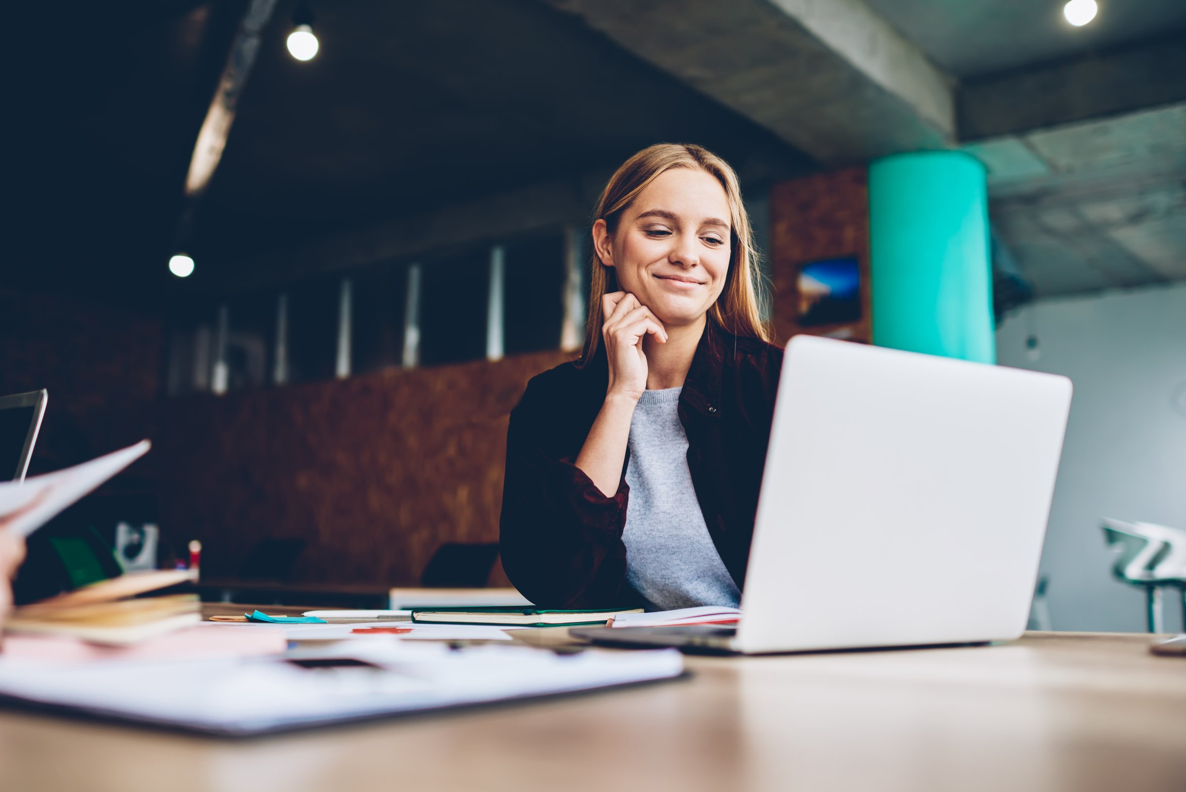 Een glimlachende vrouw leest iets op haar laptop aan een bureau vol papieren