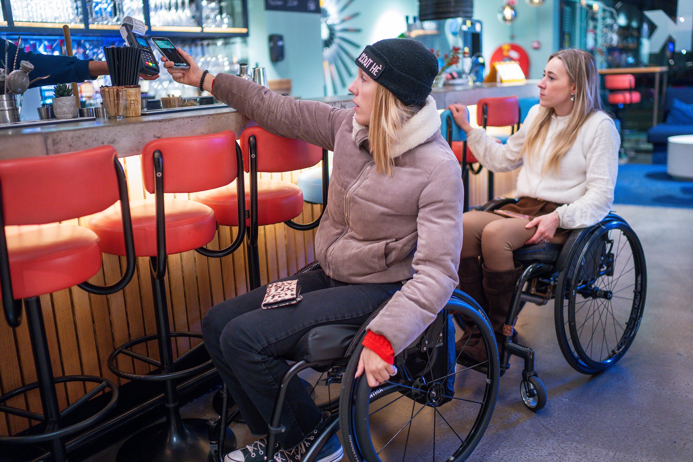Een vrouw in een rolstoel rekent aan de bar af met haar telefoon, terwijl een tweede vrouw in een rolstoel naast haar wacht.