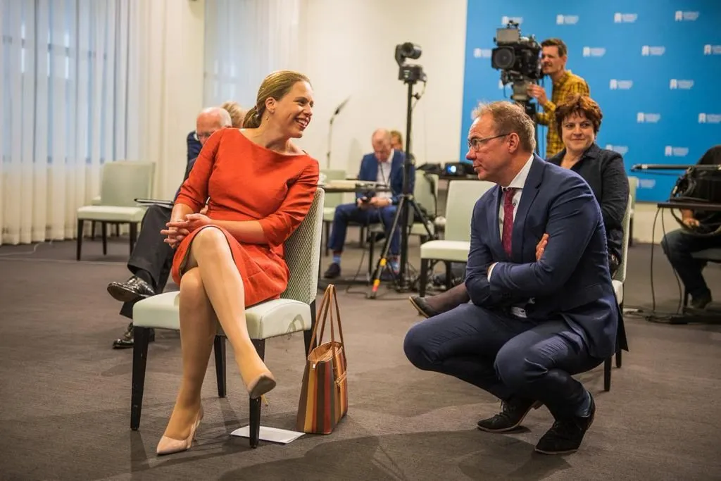 Een interview op een podium: een vrouw in een rode jurk zit op een stoel, een man in pak zit gehurkt naast haar, met publiek op de achtergrond.