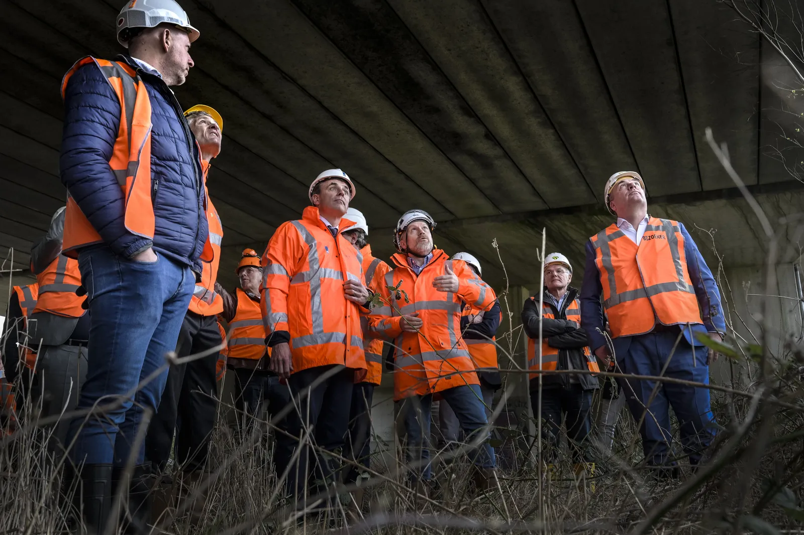 Een groep mensen in oranje veiligheidshesjes en witte helmen krijgt uitleg onder een viaduct.