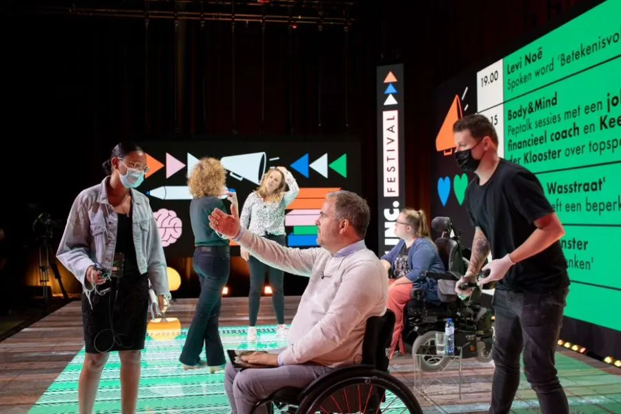 Een evenement met een kleurrijk podium; onder de aanwezigen zit iemand in een rolstoel.