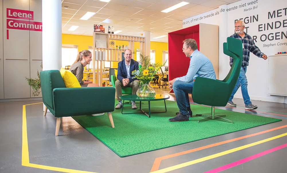 Het kantoor van Leene met groene banken en een gekleurde vloermarkering; twee mensen zitten in gesprek.