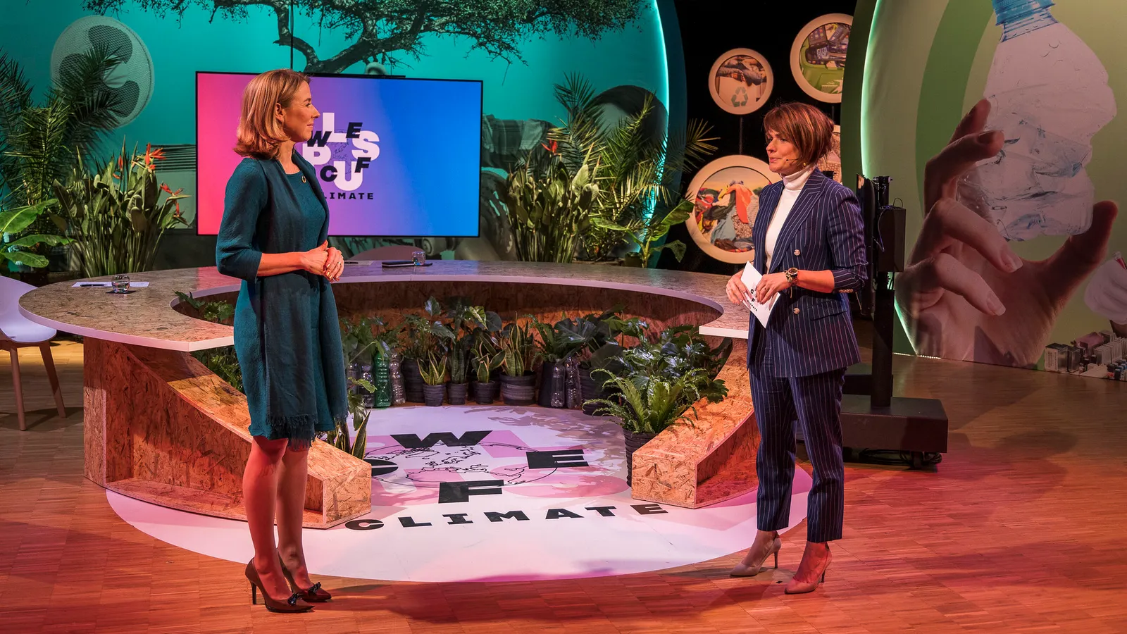 Een spreker op een podium vol planten, met een groot scherm achter zich en het woord “climate” op de vloer.